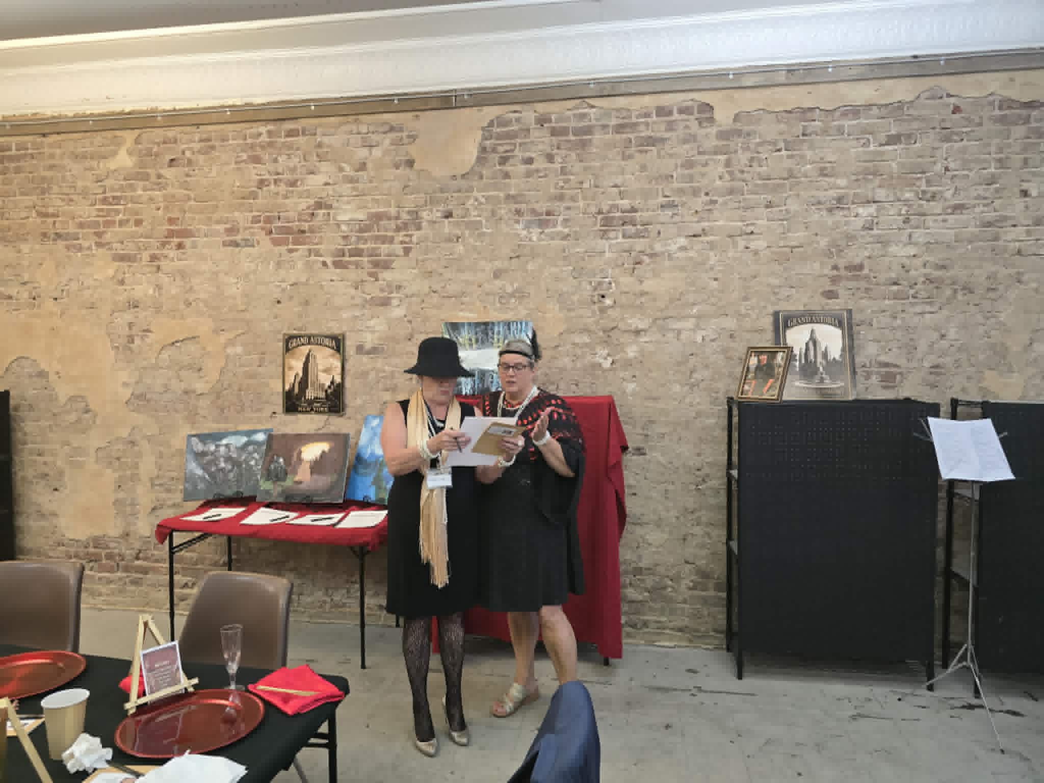 Two guests reviewing papers beside the silent-auction paintings and Grand Astoria posters.